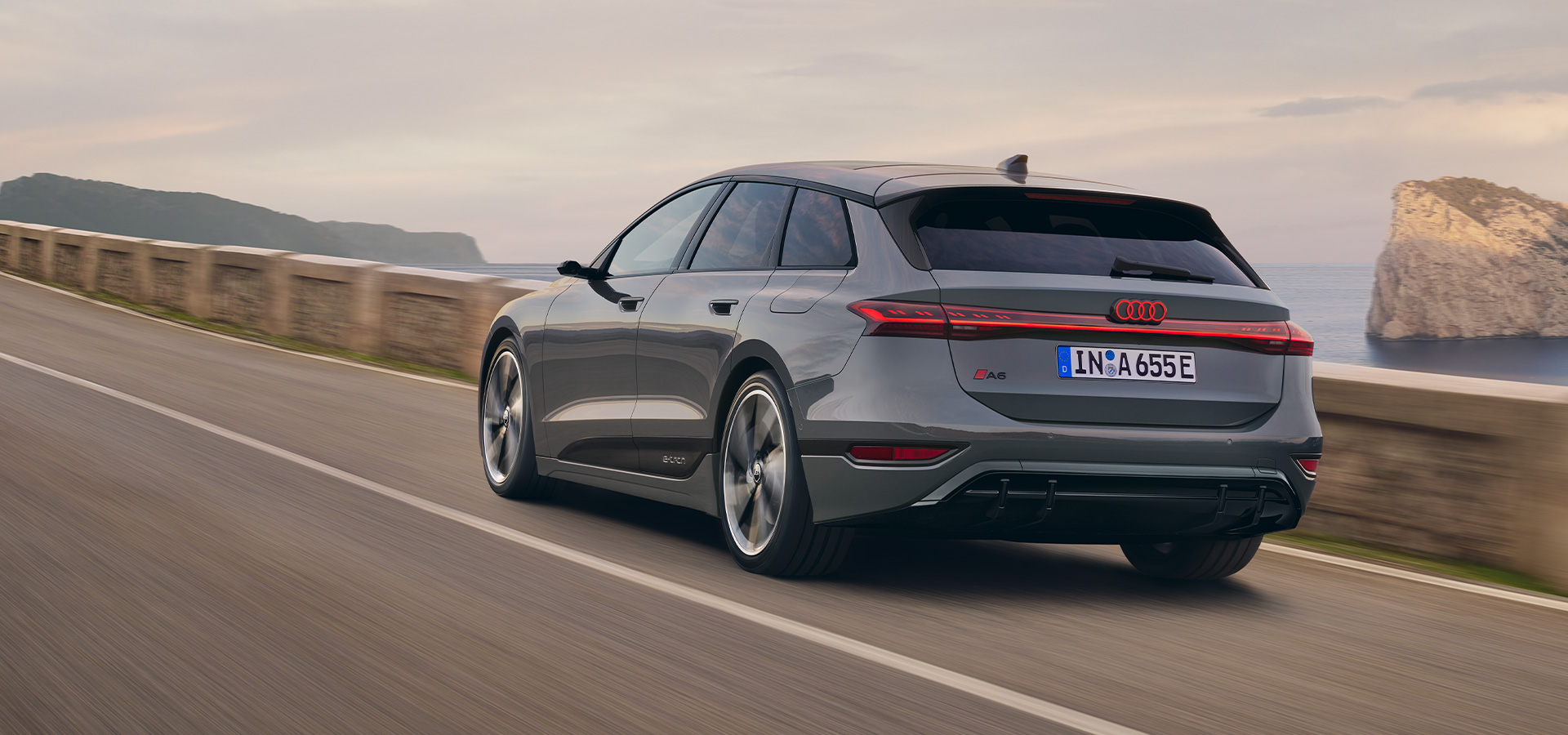 Rear view of a grey Audi A6 Avant e-tron driving along a coastal road at sunset, with aerodynamic design, LED taillights, and e-tron badging visible.