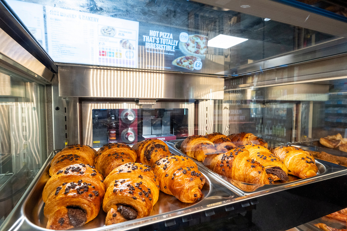 Close-up of pastry display at Co-op On the Go showing chocolate croissants under signage promoting hot pizza and bakery items