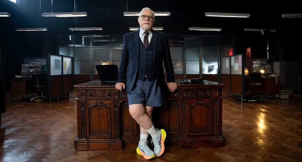 Brian Cox in the ASICS Desk Break ad poses in front of the office desk, running shoes on.