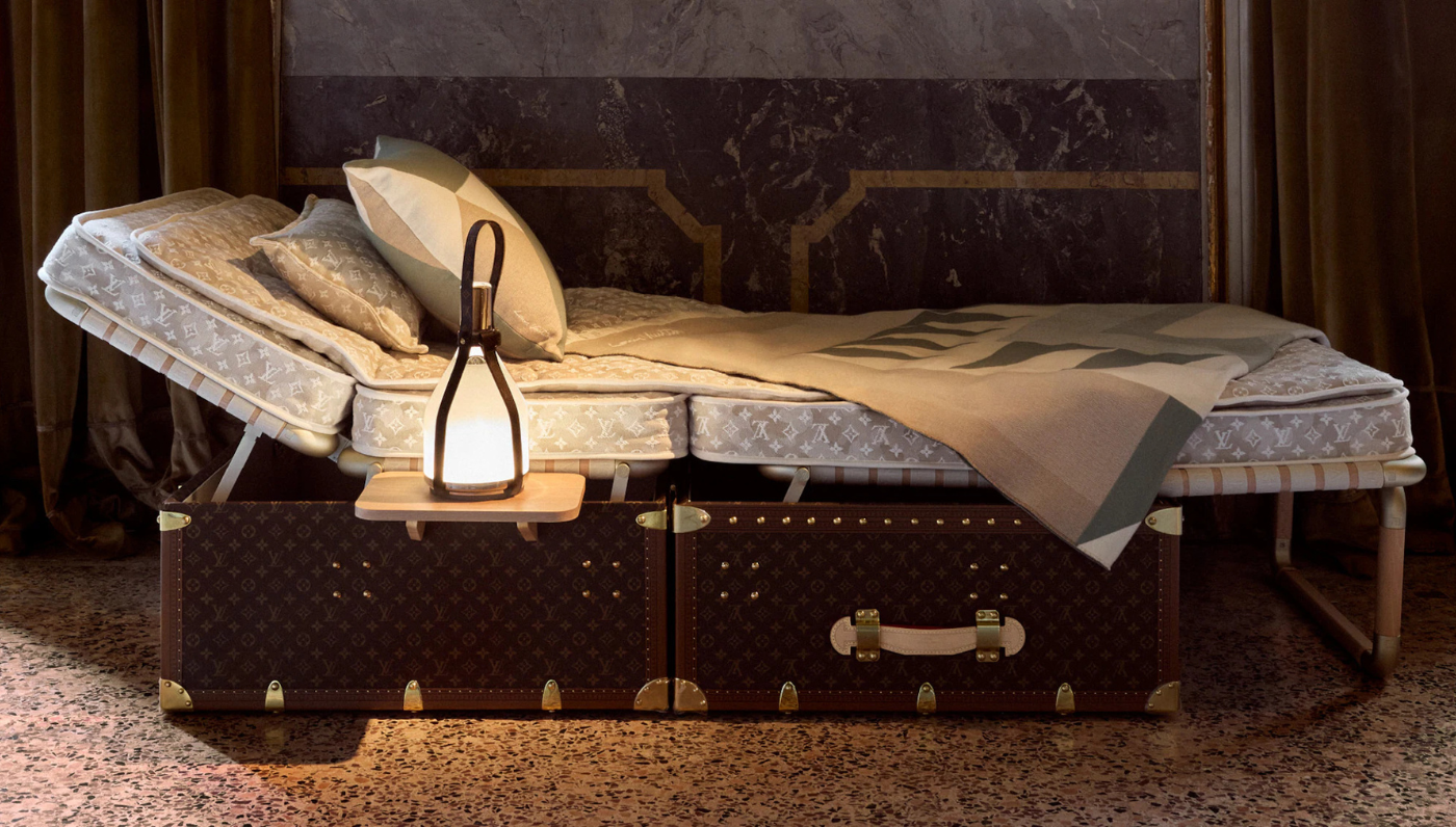 A Louis Vuitton daybed styled as a vintage travel trunk, featuring monogrammed upholstery, a sculptural bedside lantern, and a wool throw in muted earth tones—part of the maison’s new home collection presented during Milan Design Week 2025.