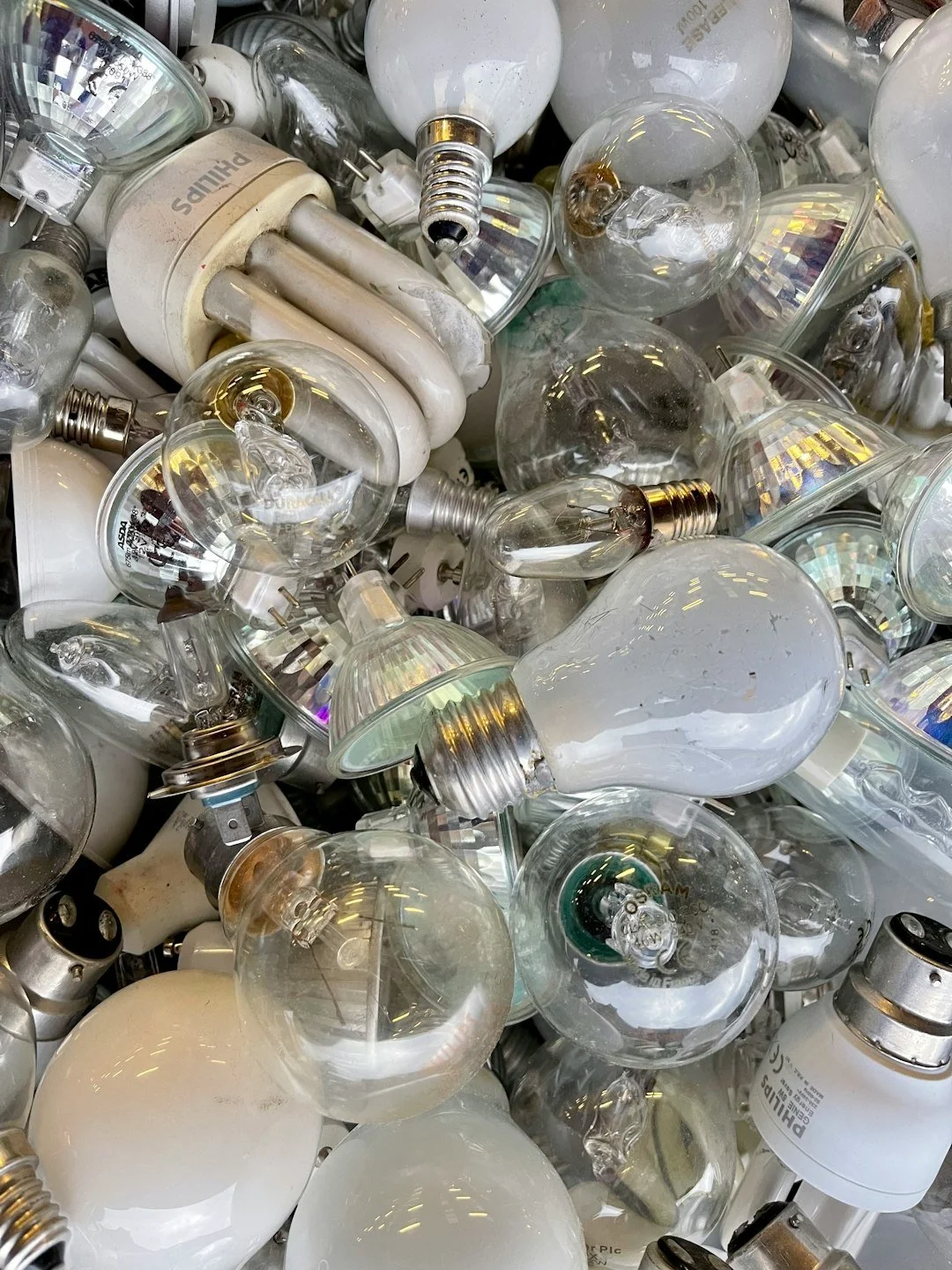 Pile of assorted used light bulbs of various types including incandescent, fluorescent, halogen, and reflector bulbs.