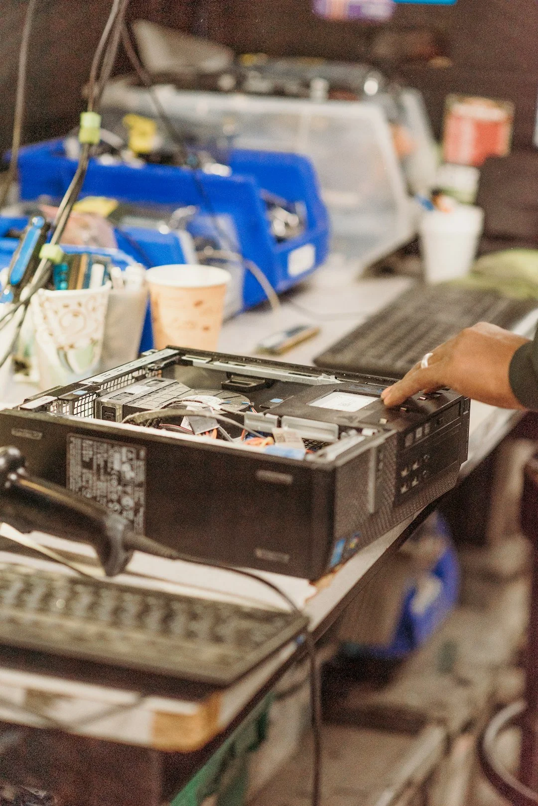 Desktop computer case lying open on a cluttered workbench with visible internal components, a hand reaching to touch it, nearby keyboard, and disorganized tools and containers in the background.