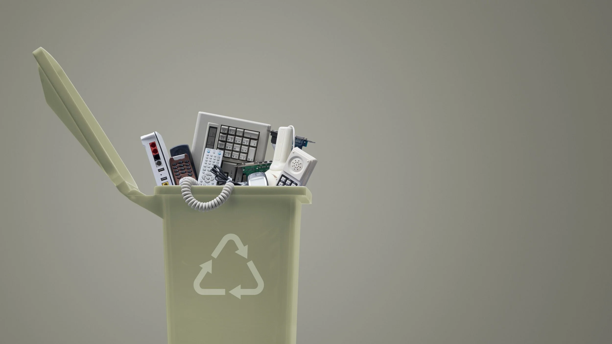 Light green recycling bin with open lid filled with various old electronic devices including remote controls, a keyboard, a mobile phone, and circuit boards, against a plain gray background.