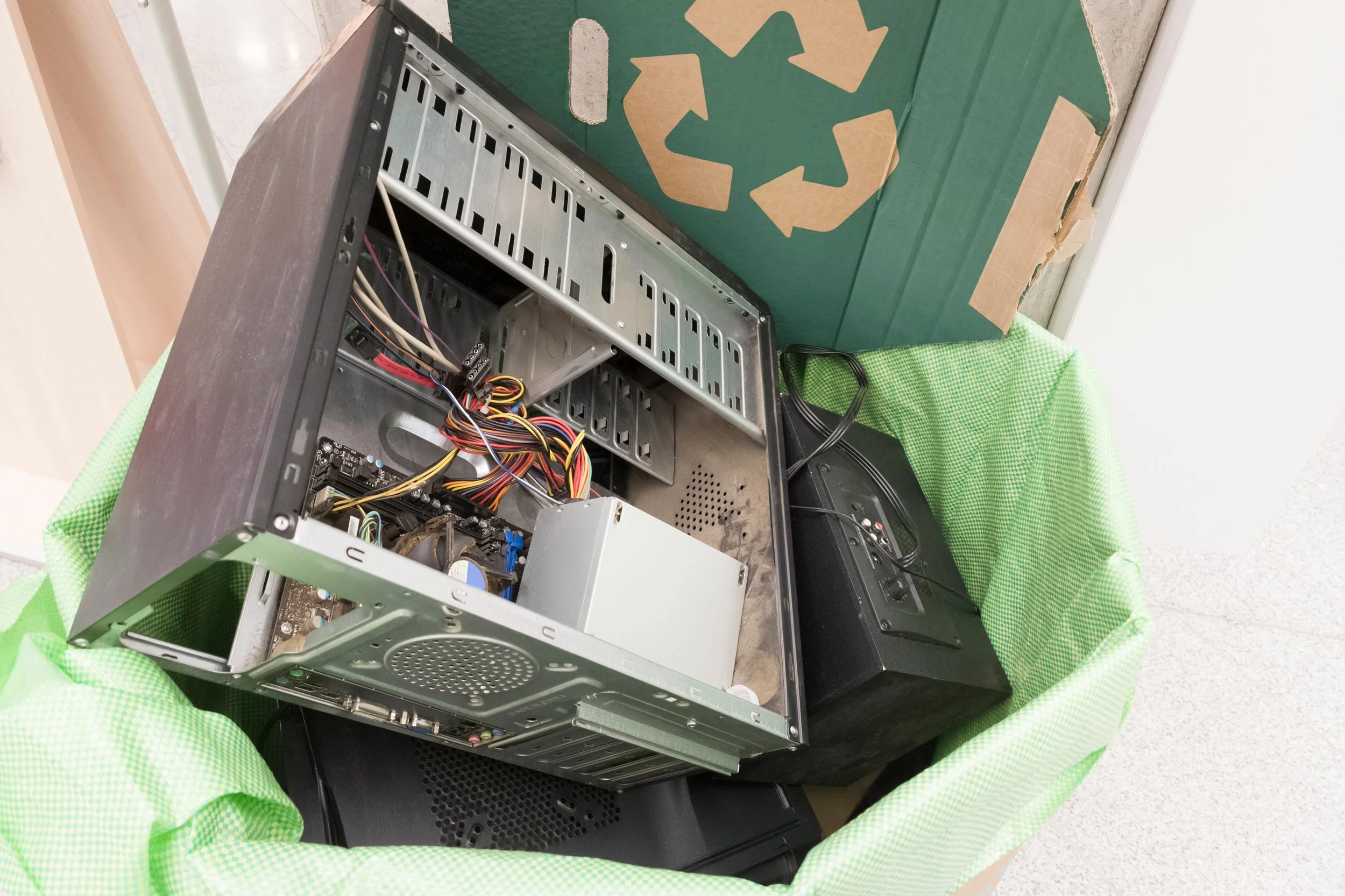 An open desktop computer case with exposed internal components and cables placed inside a green recycling bin lined with a green cloth, alongside another electronic device and a cardboard piece with a recycling symbol.