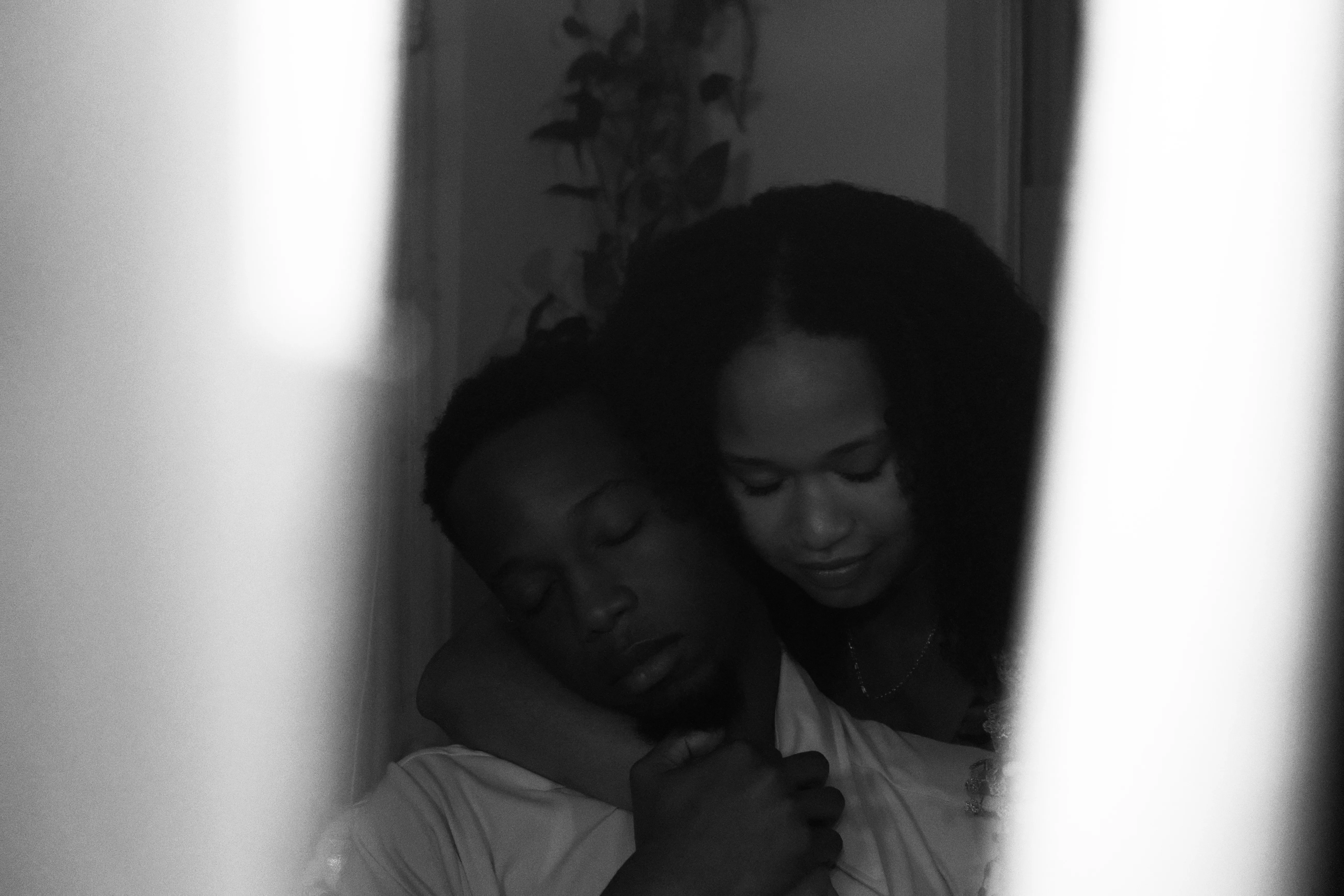 Otis rests his head against his wife as she holds him, eyes closed, seen through a narrow gap between bright panels