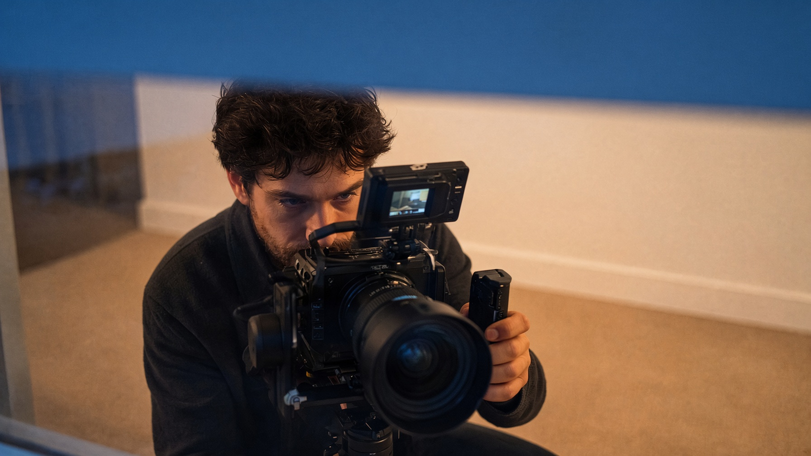 Seen through a window at dusk, a videographer inside an empty rental aims his camera at the glass.