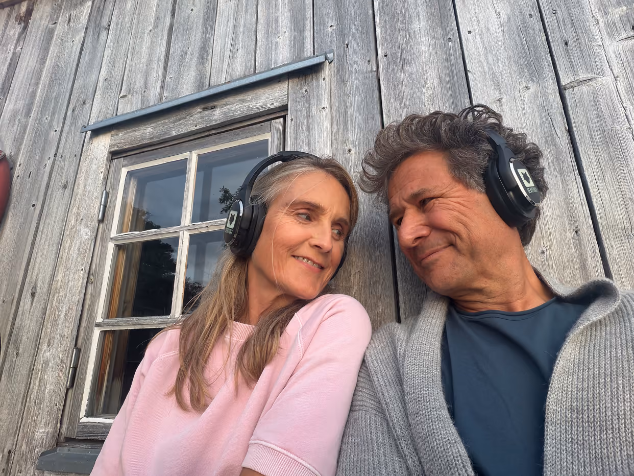 A man and woman sit closely outside a weathered wooden building with a window behind them. Both wear black over-ear headphones and look at each other smiling warmly, with the woman in a pink shirt and the man in a blue shirt and gray cardigan.