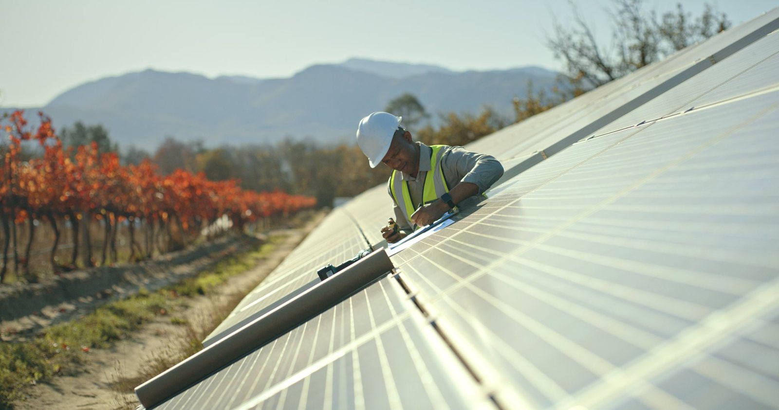 Engineer in a hard hat checking a clipboard while working on a row of solar panels