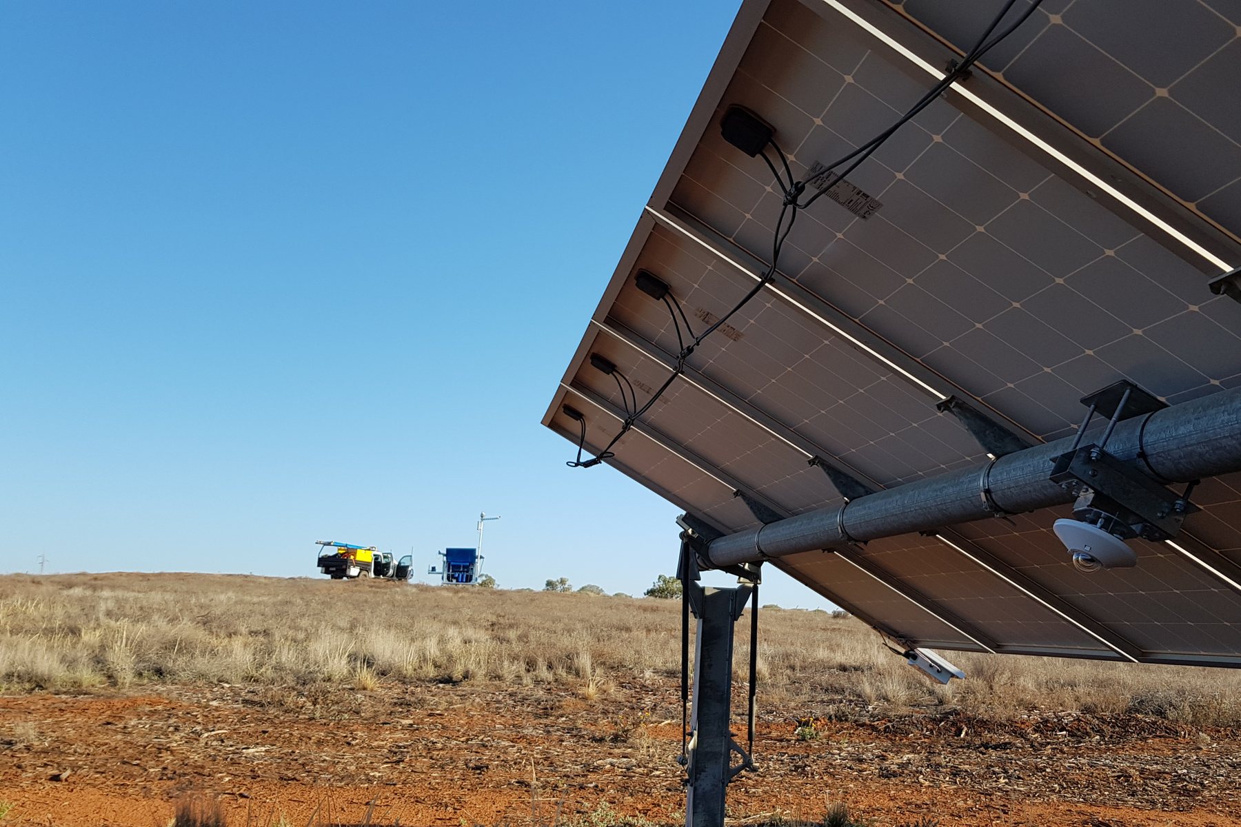 Underside of a solar tracker row with a sky camera and sensors mounted on the torque tube, a NEXO station and service vehicle in the distance