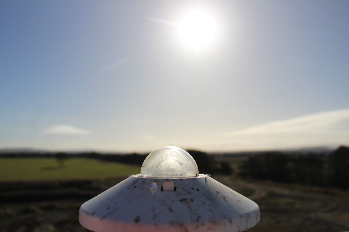 Dusty pyranometer dome on a weather station, backlit by the sun over farmland