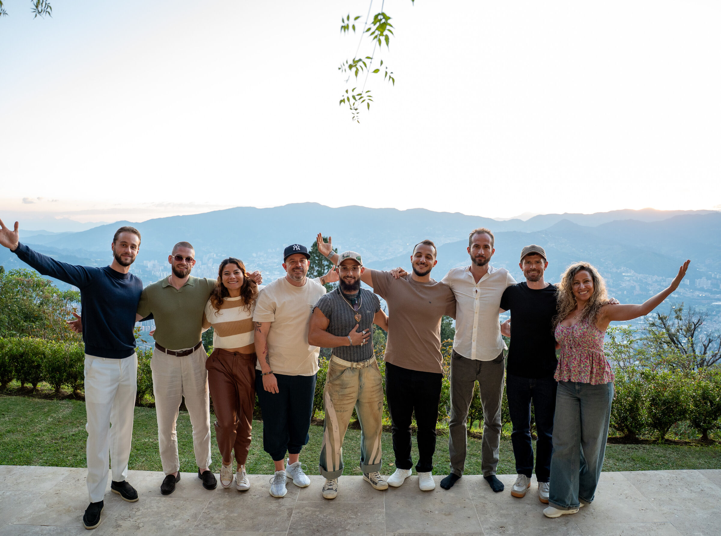 A group of THE ONE founders standing together on a hilltop terrace, arms open, mountains behind.