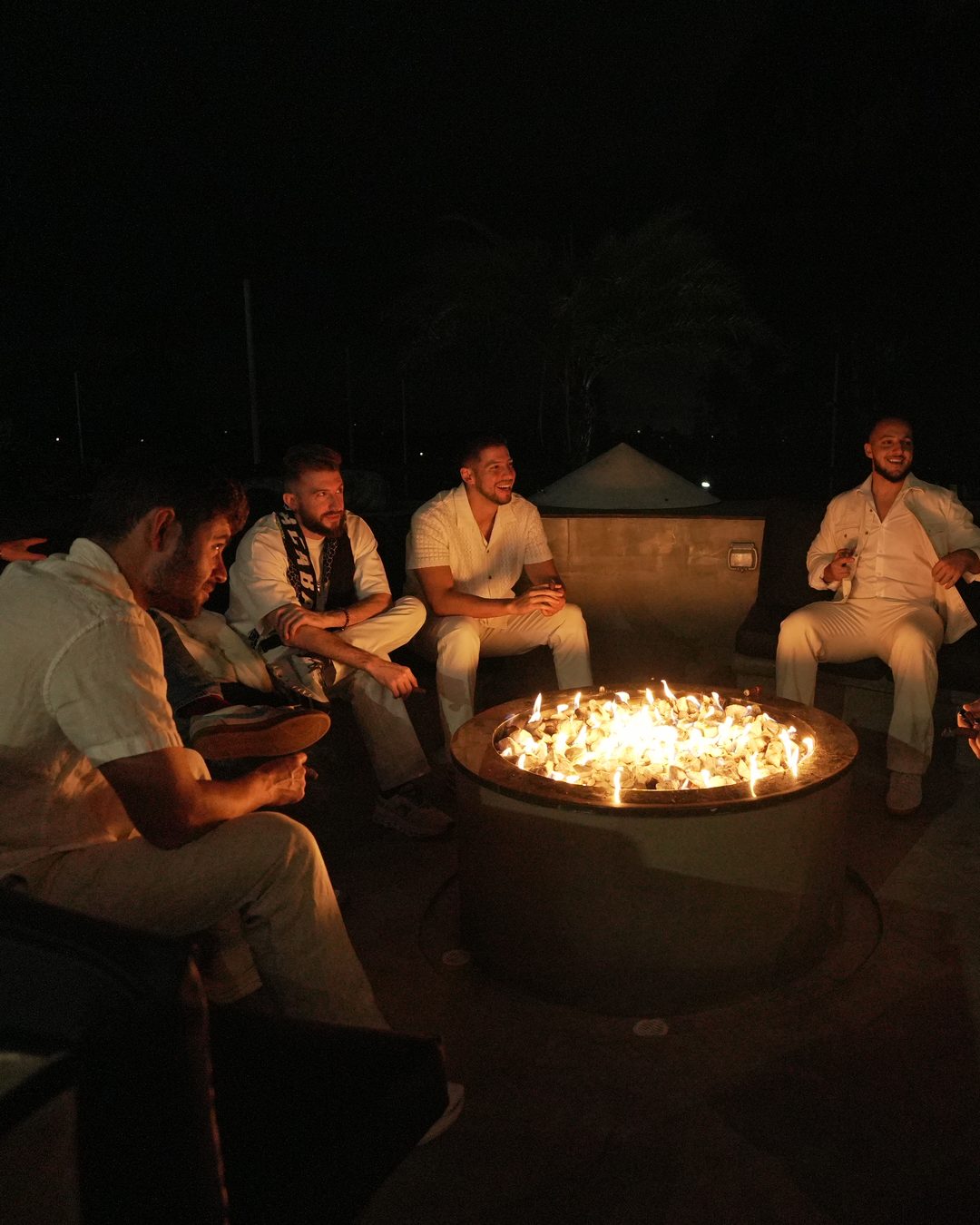 Founders Reset attendees dressed in white, seated in a circle and talking around a glowing fire pit at night.