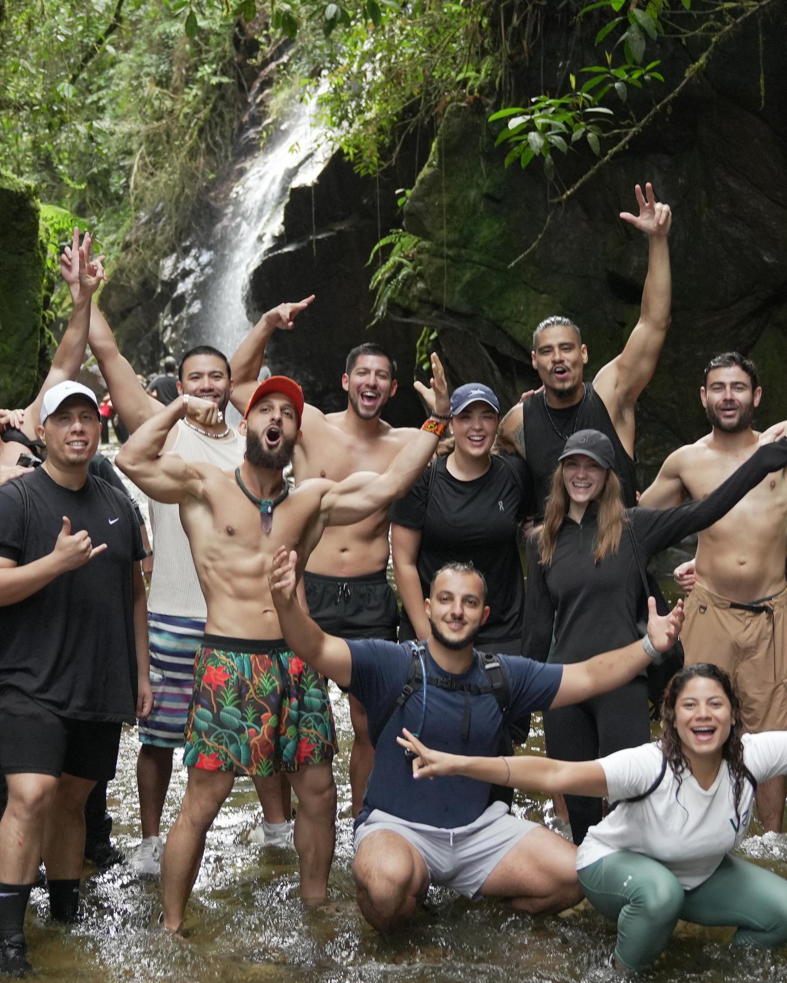 Founders Reset attendees cheering and laughing together, arms raised, in a river below a jungle waterfall on a group hike.