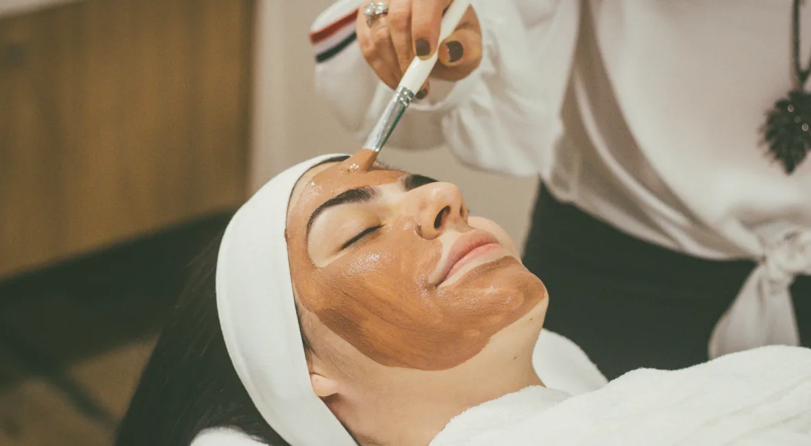 An esthetician applying a clay mask with a brush to a relaxed client wearing a white headband