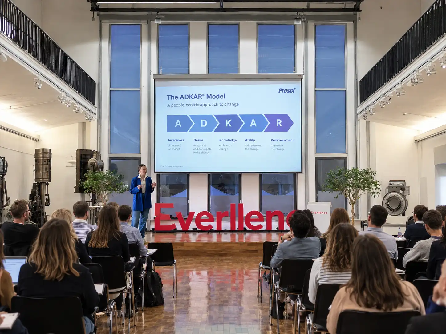 A speaker in a blue blazer presenting in front of an audience in a large room with tall windows, showing a slide about the ADKAR Model for change management on a screen.