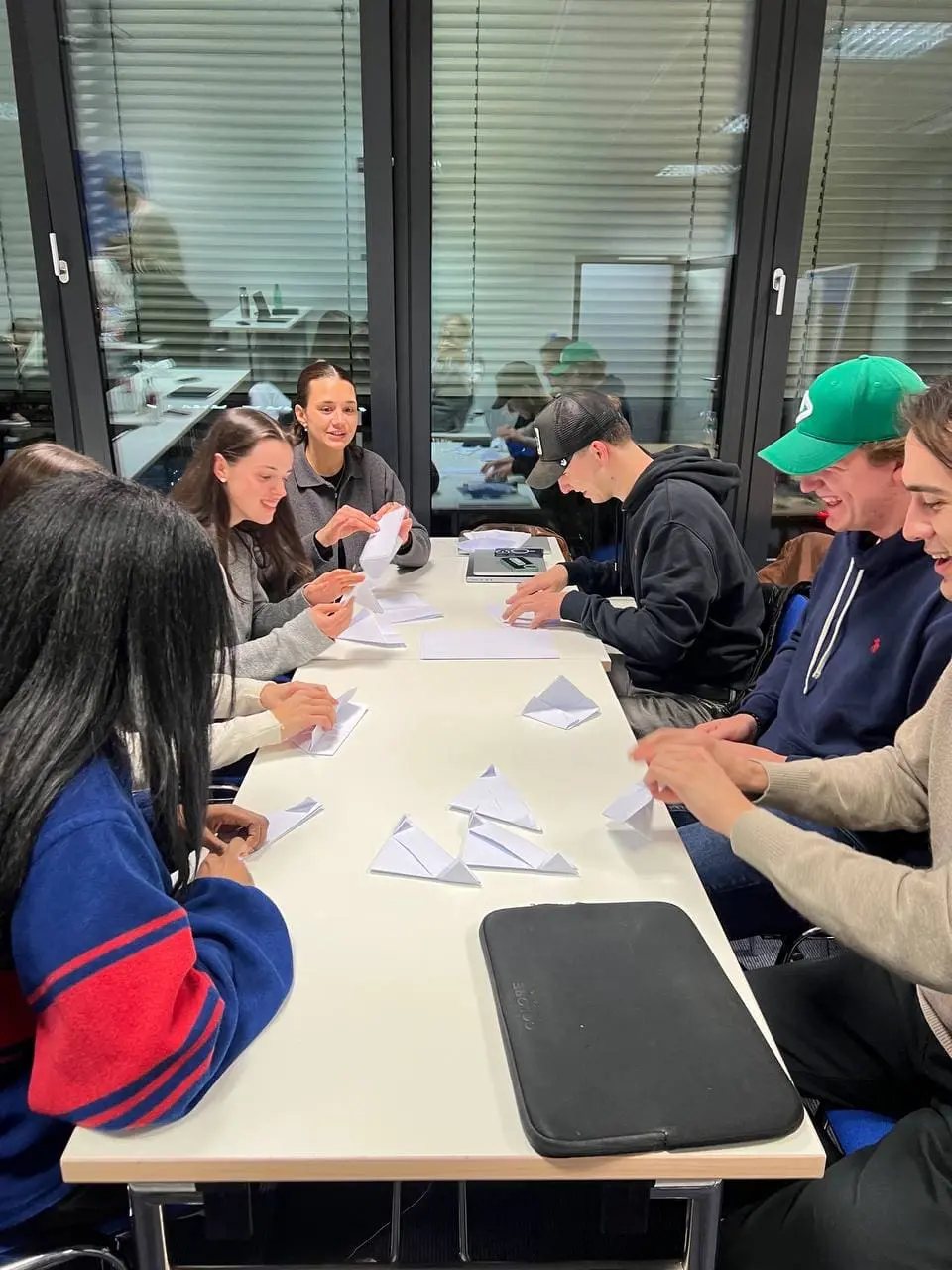 Seven young people sitting around a white table folding white paper into triangular shapes, engaging in a group activity in a room with large glass windows and blinds.