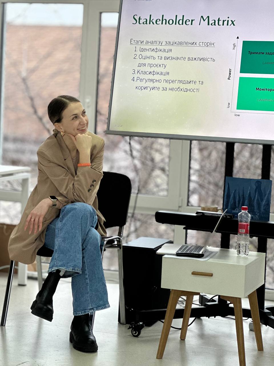 A woman in a beige blazer and blue jeans sits on a chair smiling, next to a large screen displaying a stakeholder matrix with text in Ukrainian and a chart showing power versus interest levels.