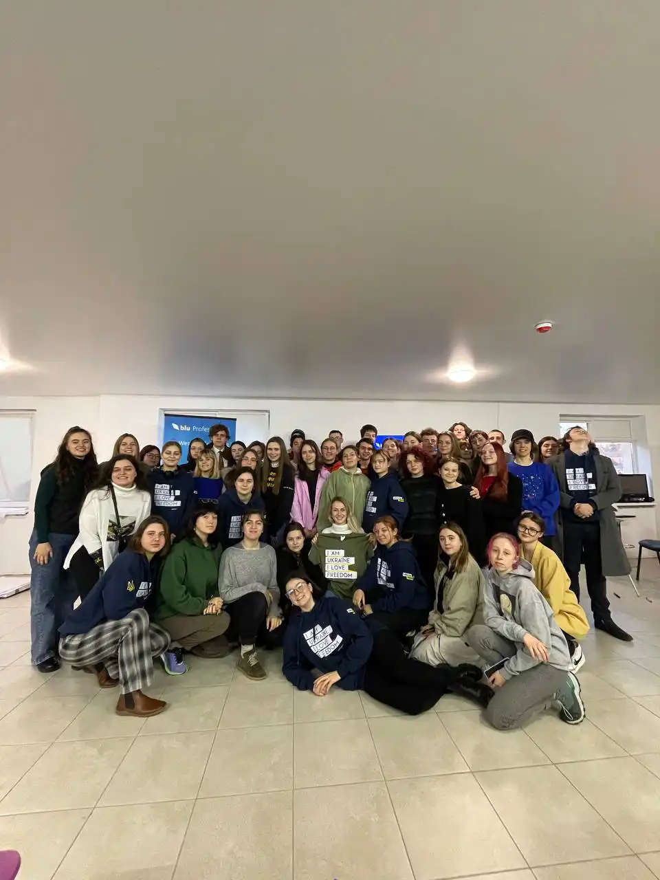 A large group of young adults posing together indoors on a tiled floor under bright ceiling lights, some wearing navy hoodies with white text reading 'I AM UKRAINE I LOVE FREEDOM.'