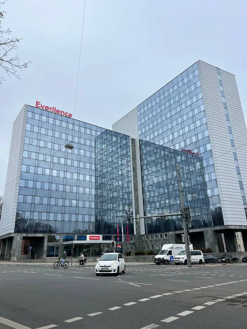 Modern glass office building with 'Everlence' signage on top and above the entrance, located at an urban intersection with vehicles and cyclists on the road.