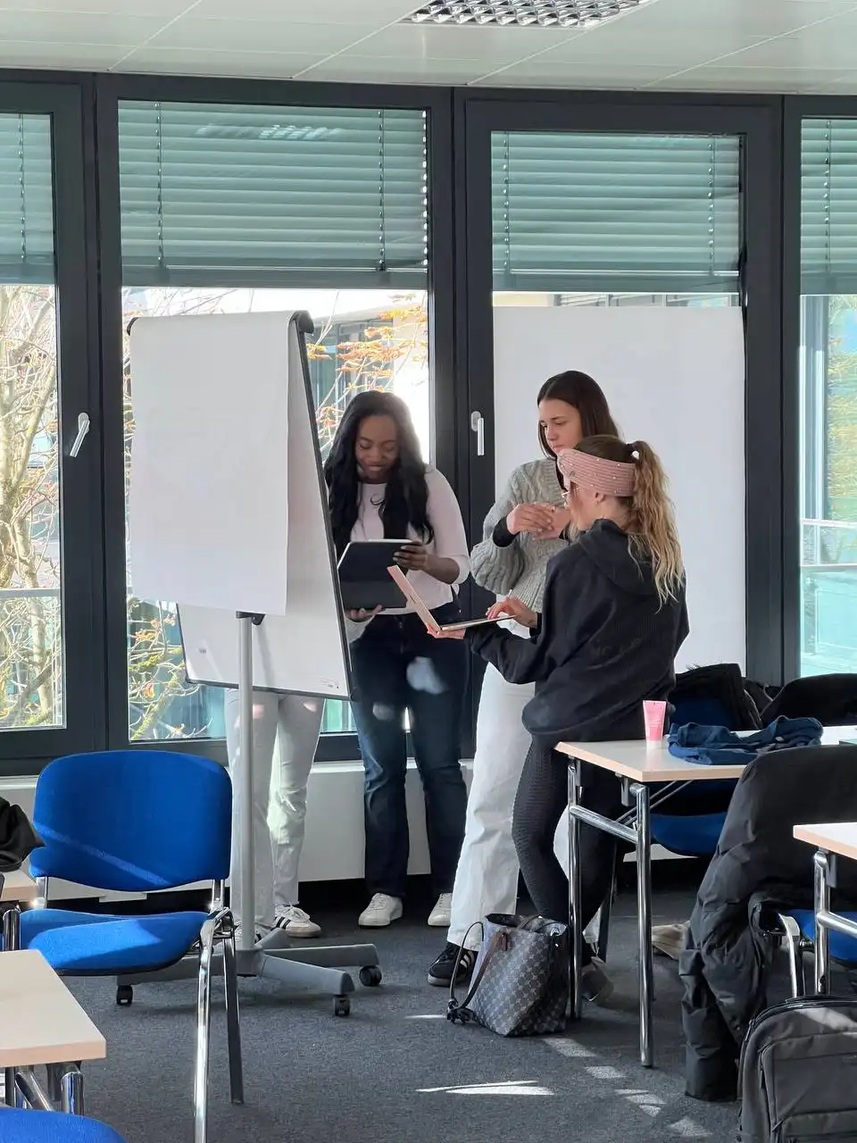 Three women standing in a modern office or classroom near large windows with closed blinds, looking at laptops and tablets while discussing, surrounded by blue chairs and tables.