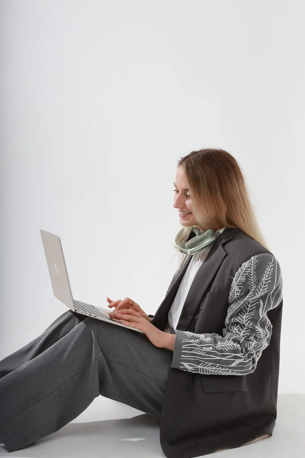 Smiling woman with long blonde hair sitting on the floor using a laptop, wearing dark gray wide-leg pants and a dark blazer with embroidered white leaf patterns on the sleeves, and headphones around her neck.