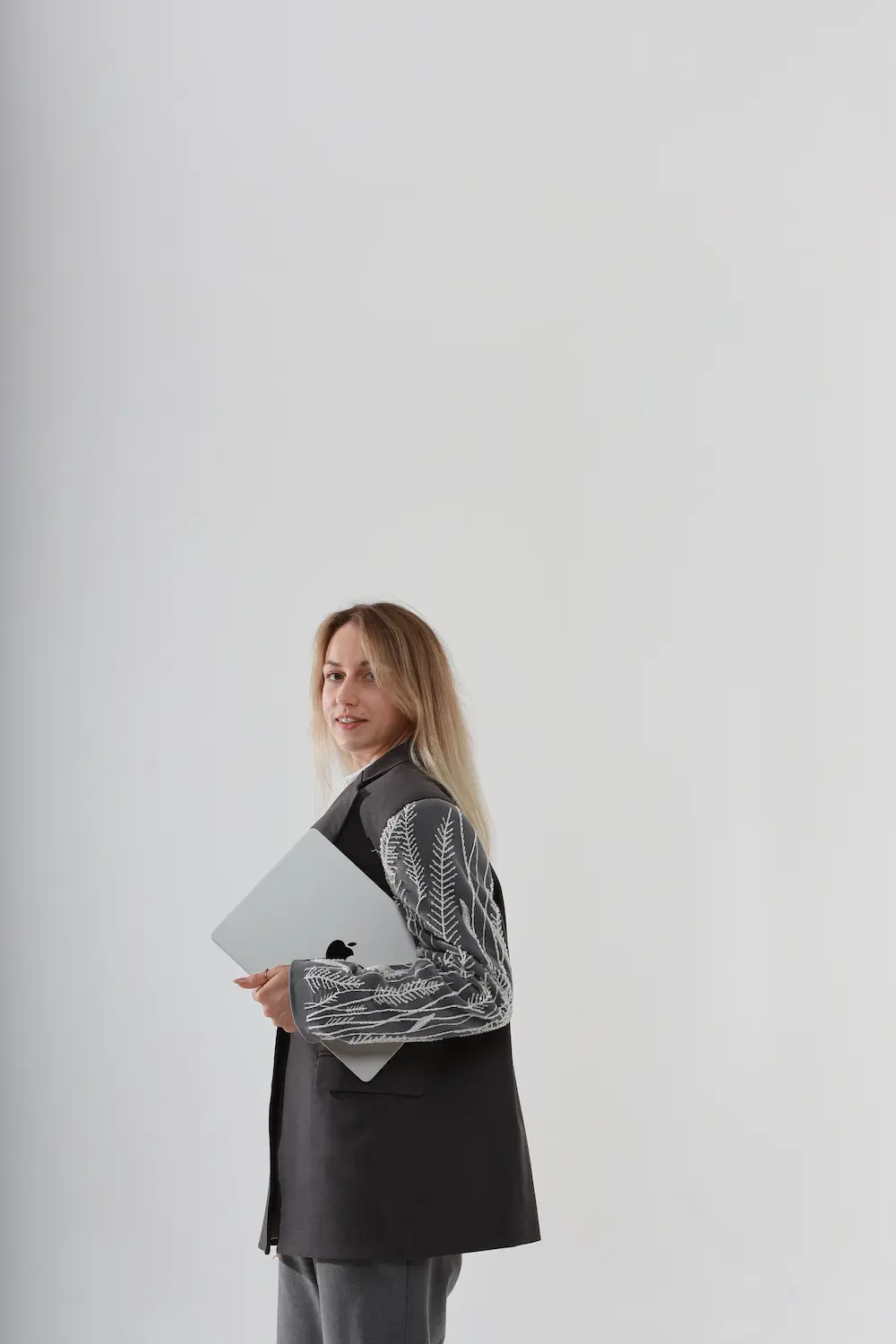 Blonde woman in a black blazer with white fern patterns on sleeves and gray pants holding a closed silver Apple laptop, standing against a plain light gray background and looking at the camera.