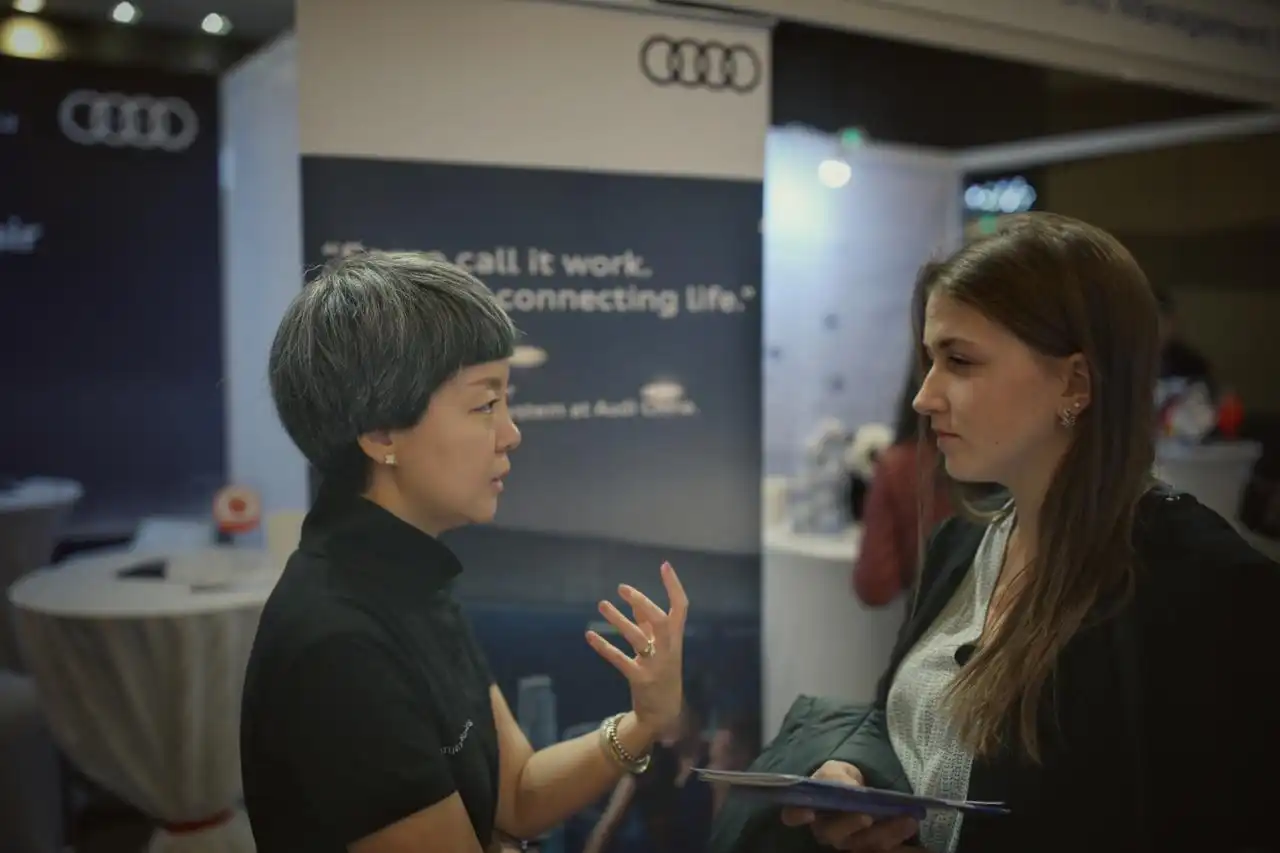 Two women engaged in a conversation at an indoor event, with one woman gesturing while speaking and the other holding a notebook and listening attentively, Audi logos visible in the background.