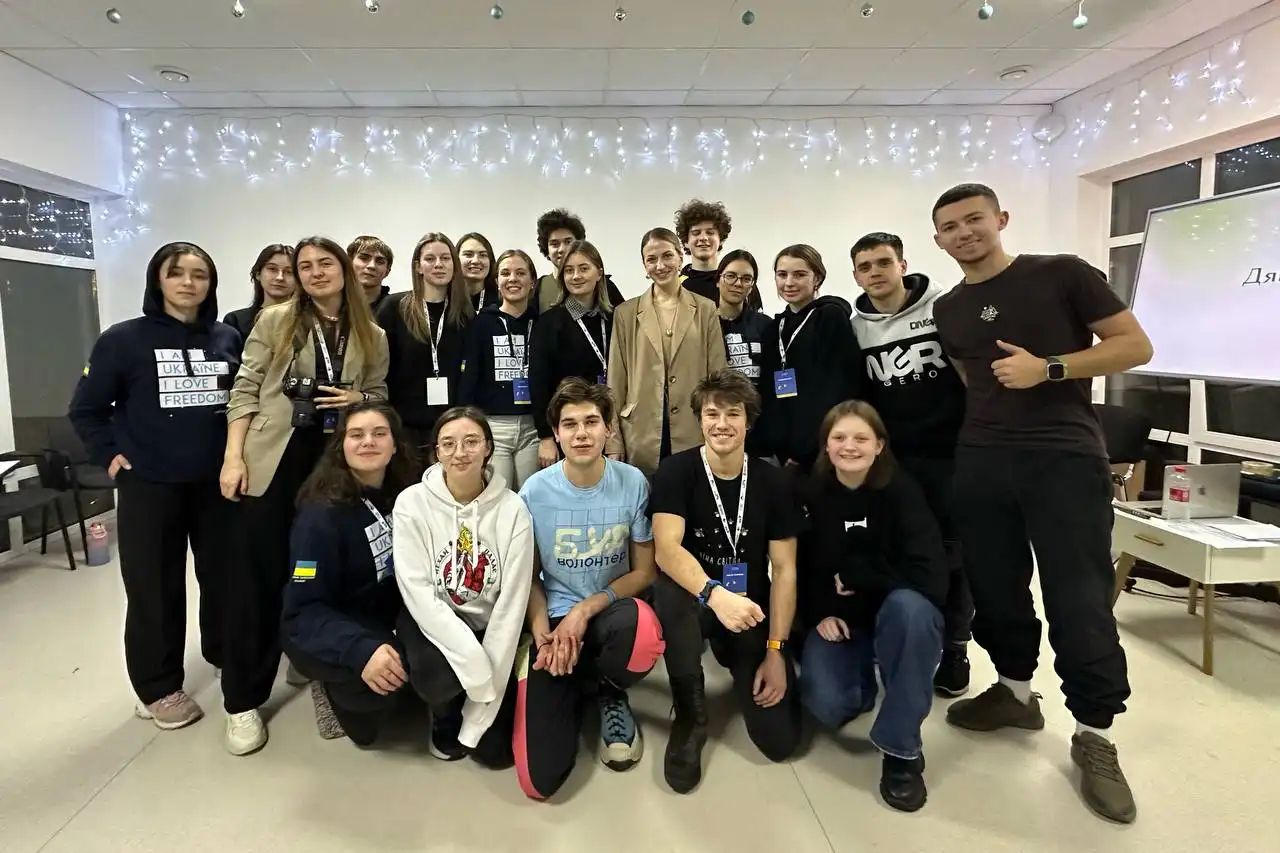 Group of young people in a well-lit room posing for a photo, some wearing lanyards and hoodies with Ukrainian-themed messages, festive string lights hanging above.