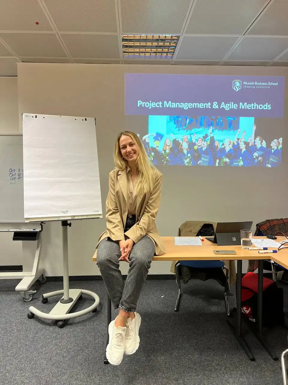 A smiling young woman with long blonde hair wearing a beige blazer and gray pants sits on a desk in a classroom with a blank flip chart to her left and a projected presentation titled 'Project Management & Agile Methods' from Munich Business School on the wall behind her.