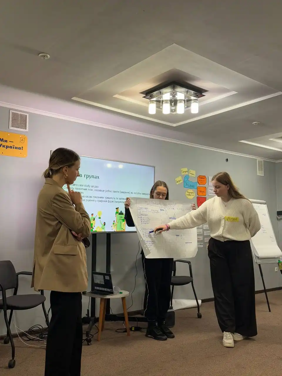 Three women in a conference room; one woman stands on the left in a tan blazer observing, one in the center holds a large sheet of paper with notes and charts, and the third woman on the right points to the paper with a pen. Behind them is a TV screen displaying text in Ukrainian and colorful sticky notes on the wall.