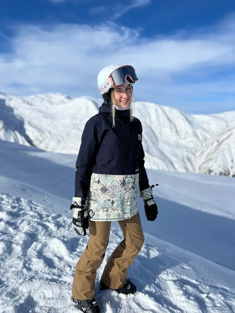 Smiling person dressed in ski gear with white helmet, goggles, black and patterned jacket, brown pants, and gloves standing on snowy mountain slope with snow-covered mountains and blue sky in the background.