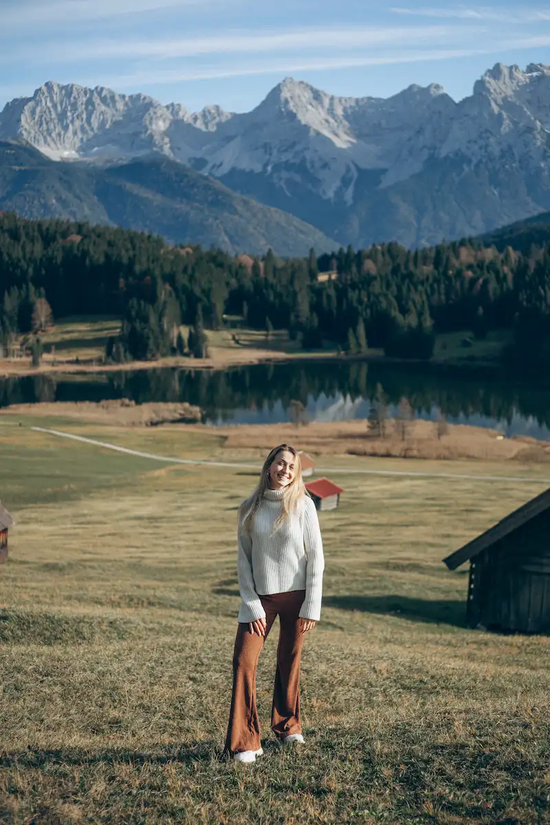 A woman in a white turtleneck sweater and brown pants standing on a grassy hill with a scenic view of a lake, forest, and snow-capped mountains in the background under a partly cloudy sky.