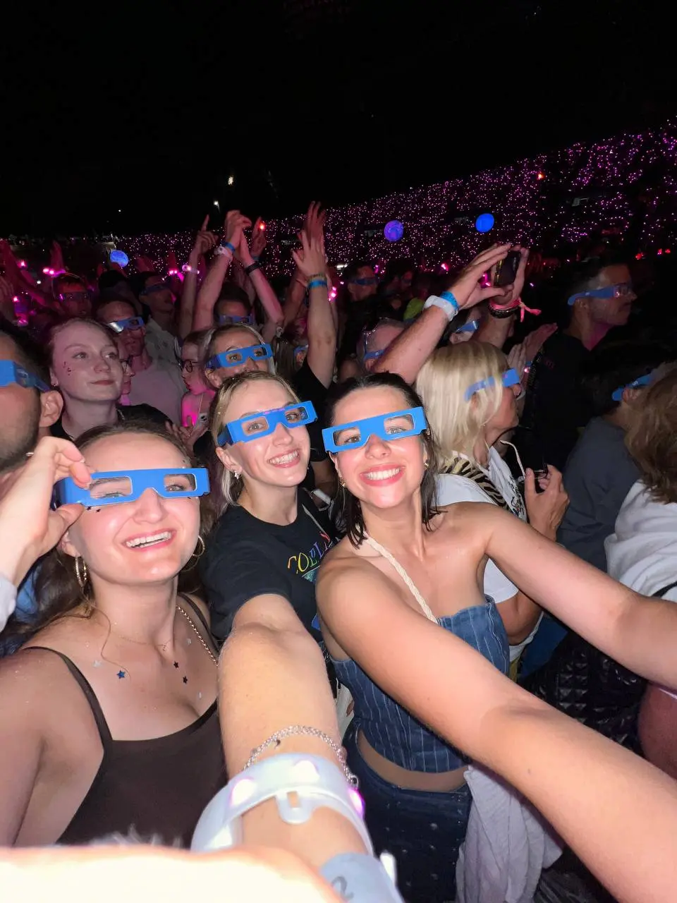 Three young women smiling and taking a selfie at a crowded nighttime event, all wearing blue 3D glasses, with a large audience and many small pink lights in the background.