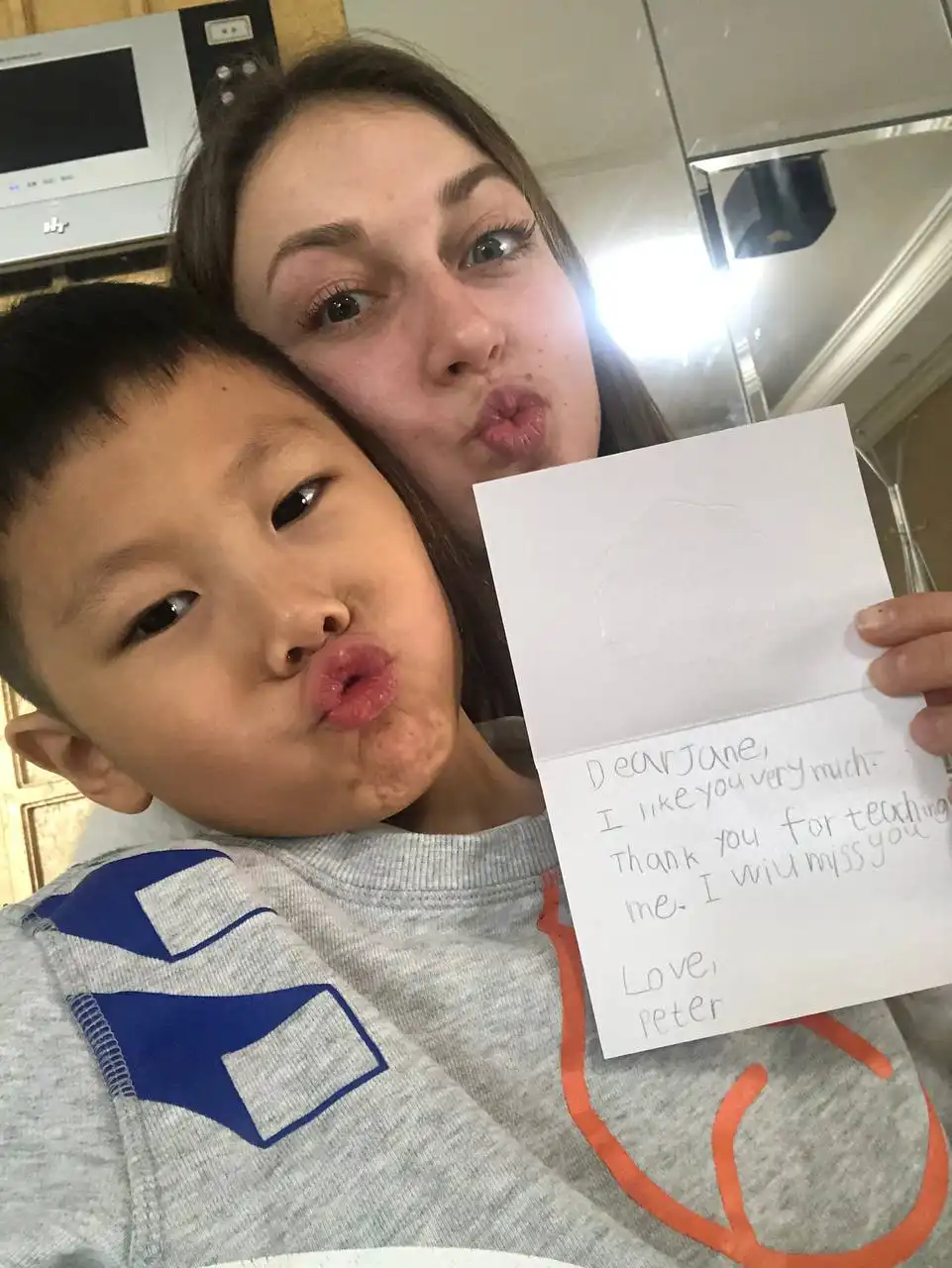 A woman and a young boy making kiss faces at the camera; the woman holds a handwritten note saying, 'Dear Jane, I like you very much. Thank you for teaching me. I will miss you. Love, Peter.'