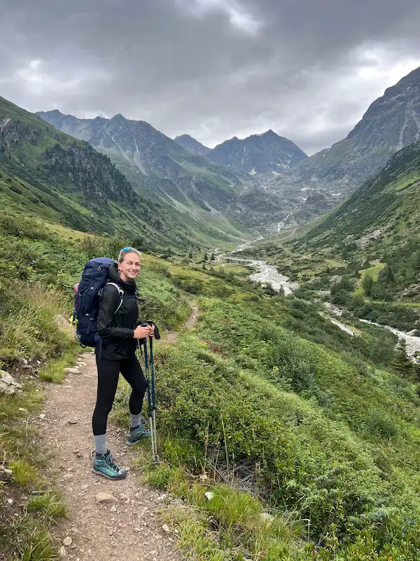 Young woman wearing hiking gear and a large backpack stands on a narrow mountain trail surrounded by green shrubs, with tall rugged mountains and a cloudy sky in the background.