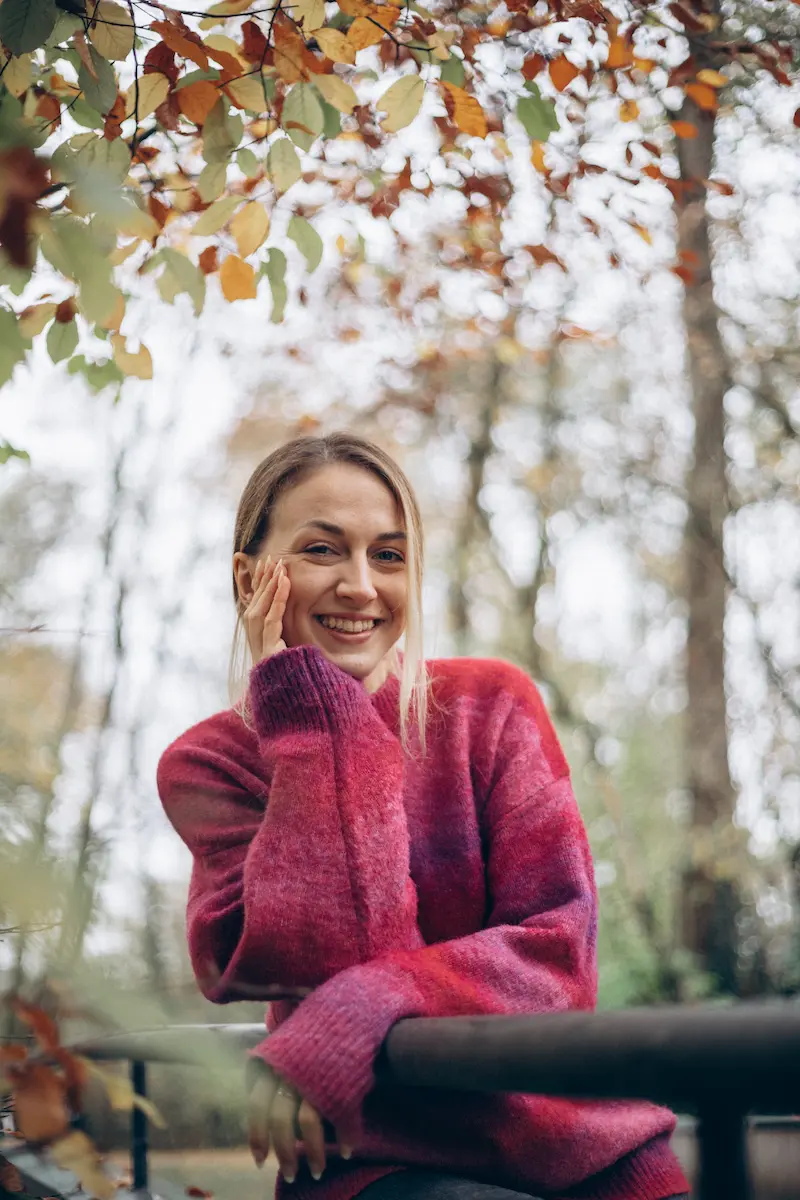 Smiling woman wearing a pink and purple sweater leaning on a railing outdoors with autumn leaves and blurred trees in the background.