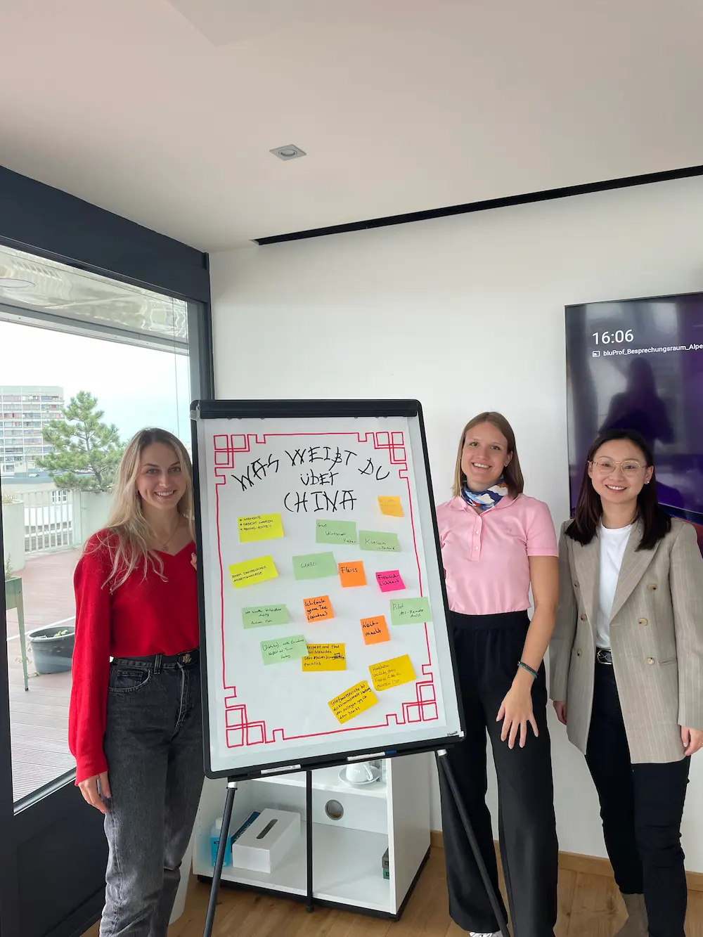 Three women standing indoors around a whiteboard on an easel with colorful sticky notes and the German phrase 'Was weißt du über China' written on it, in a modern room with a large window and a TV screen showing the time 16:06.