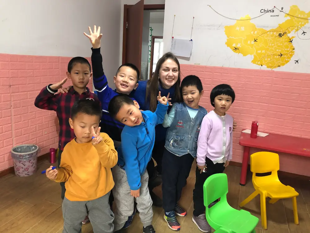 Smiling woman surrounded by six children in a classroom with pink walls, a map of China on the wall, and colorful chairs.