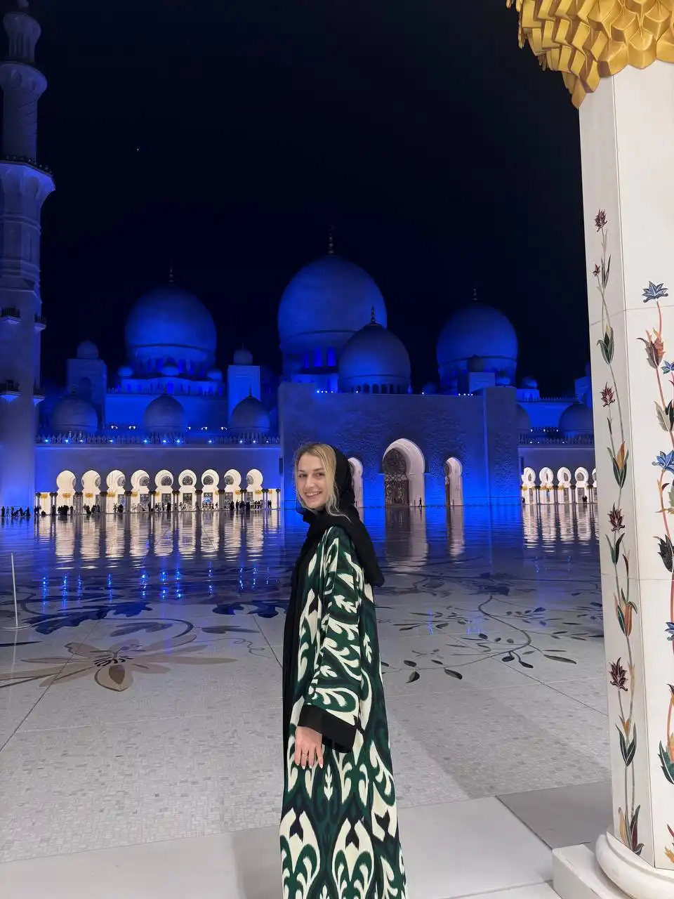 A woman wearing a black headscarf and a patterned green and white abaya stands and smiles in the foreground at night, with the illuminated blue domes, arches, and intricate floor designs of the Sheikh Zayed Grand Mosque in Abu Dhabi behind her.