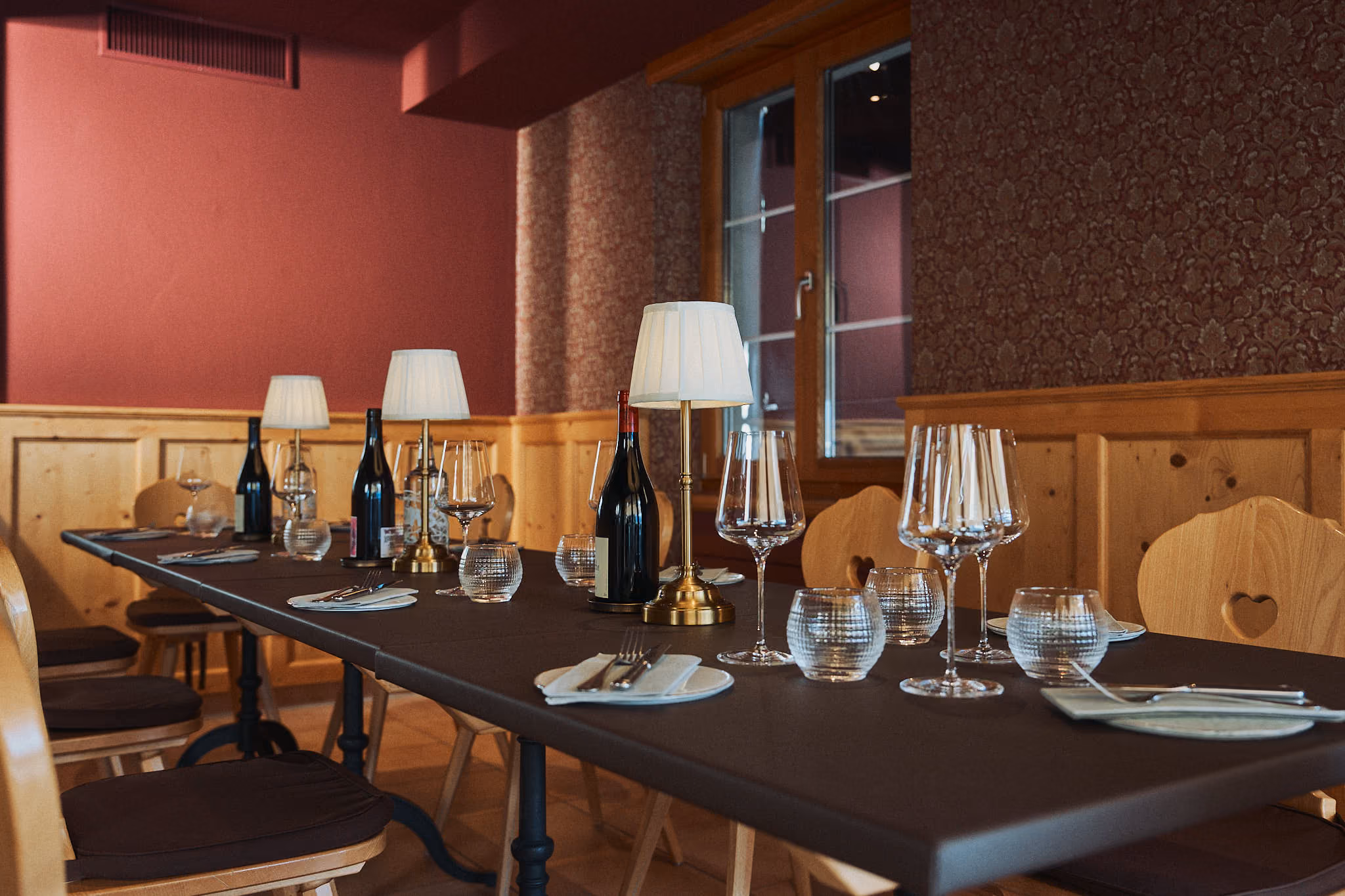 Gasthaus-Esstisch, dekoriert mit Weinflaschen, Weingläsern, kleinen Wassergläsern und zwei kleinen Tischlampen in einem Raum mit dunklen Wandverkleidungen und Holzstühlen mit herzförmiger Aussparung.