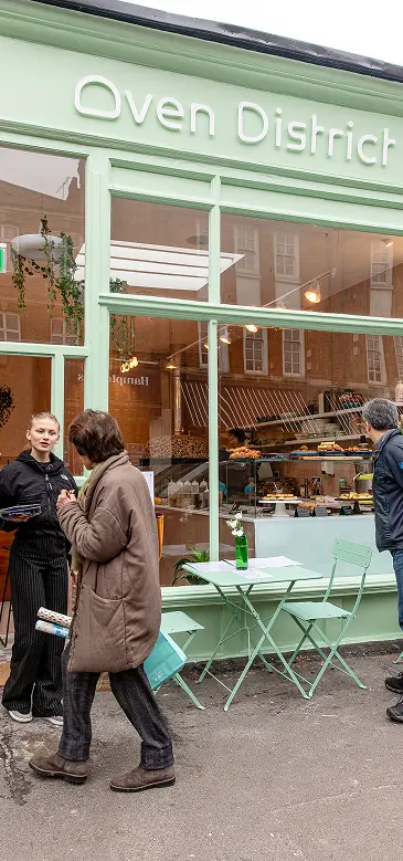 People outside Oven District bakery, beside mint-green windows and pavement tables.