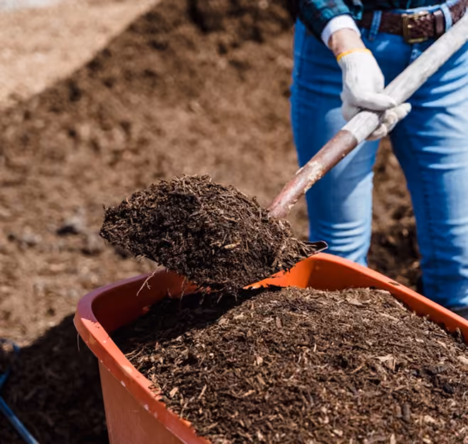 Osoba u belim rukavicama lopatom utovaruje tamnu zemlju u narandžaste kolica u spoljnjem prostoru. U prvom planu je rastresit kompost, a u pozadini se vide plave farmerke i zemljana podloga, uz jaku dnevnu svetlost.