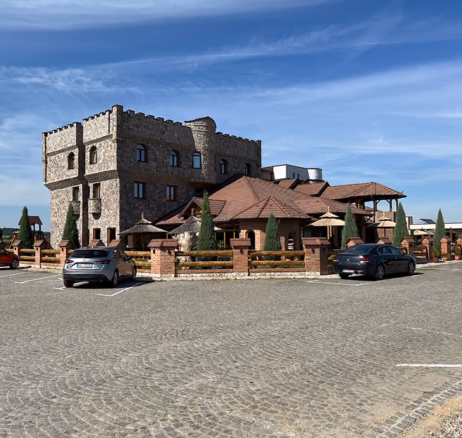 Kamena zgrada sa kulom i crvenim krovovima stoji iza niskog restoranskog kompleksa sa drvenom ogradom i čempresima. U prvom planu je širok popločan parking sa dva automobila, pod vedrim plavim nebom i jakim dnevnim svetlom.