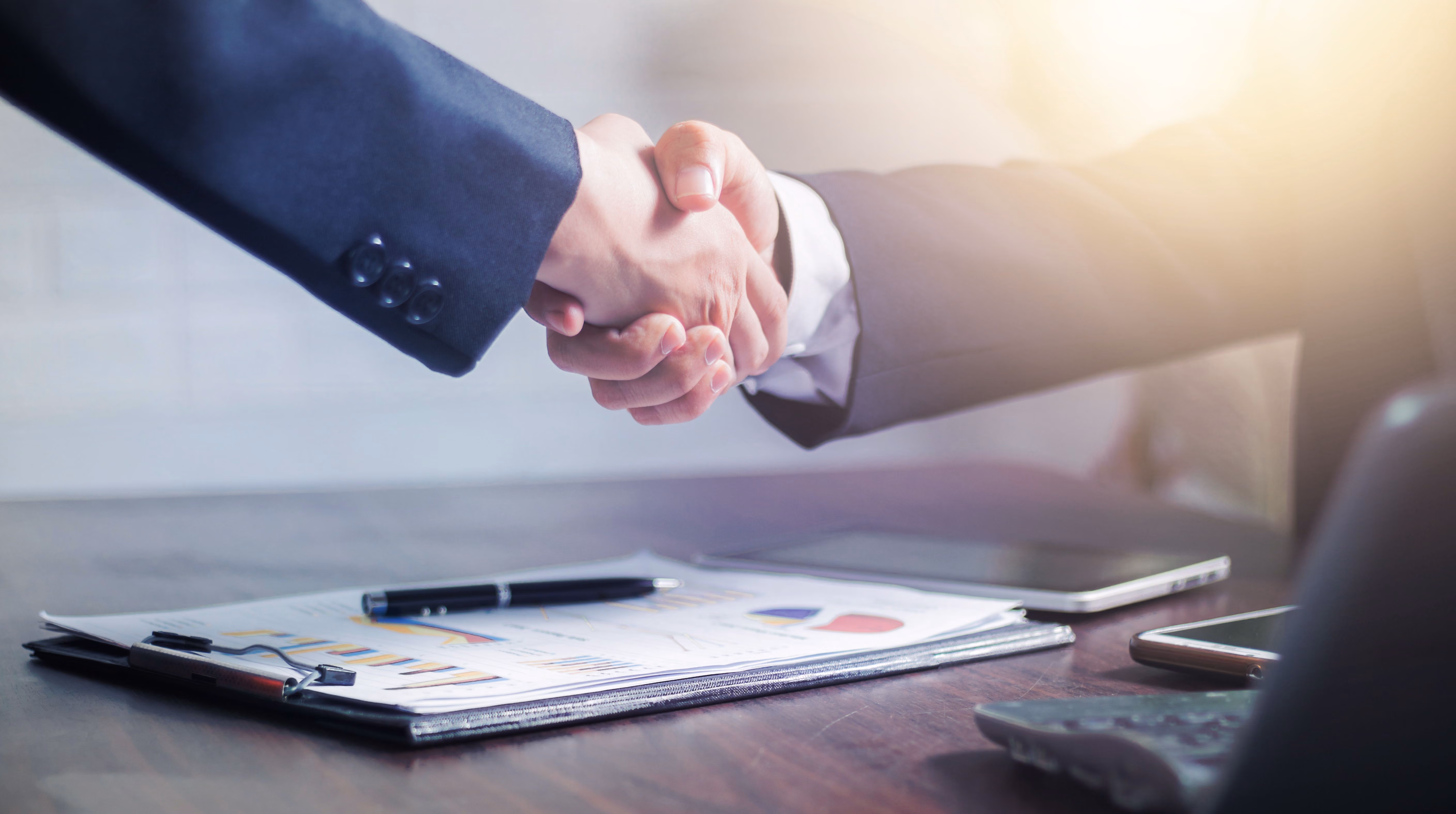 Two people in business suits shaking hands over a desk with documents, a pen, and electronic devices, with soft sunlight in the background.