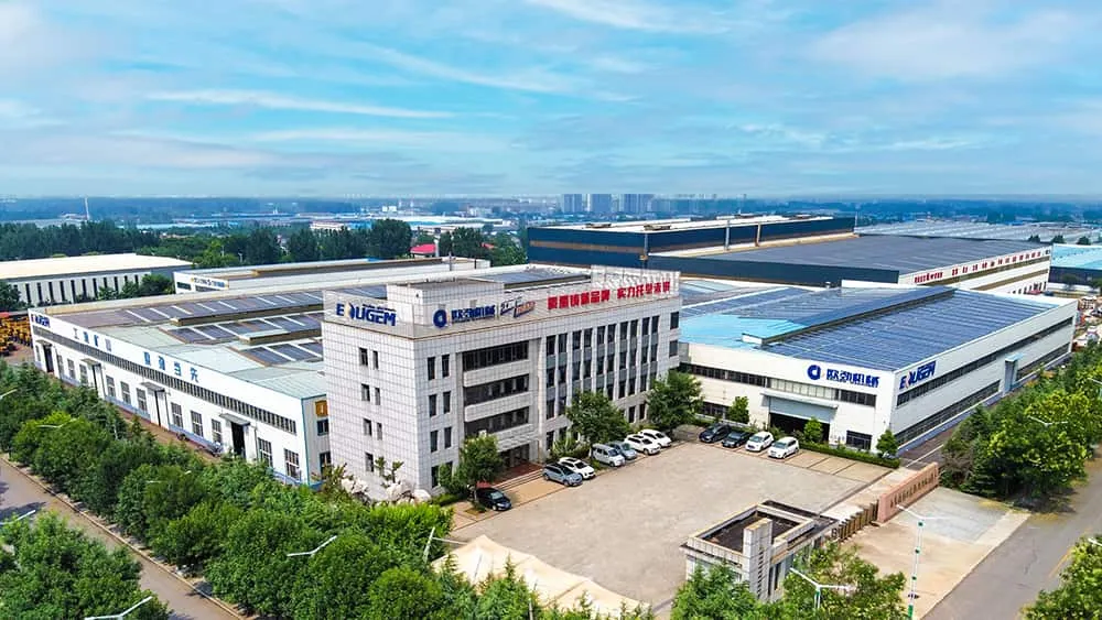 Aerial view of a large industrial factory complex with multiple white buildings featuring company logos, surrounded by green trees and clear blue sky.