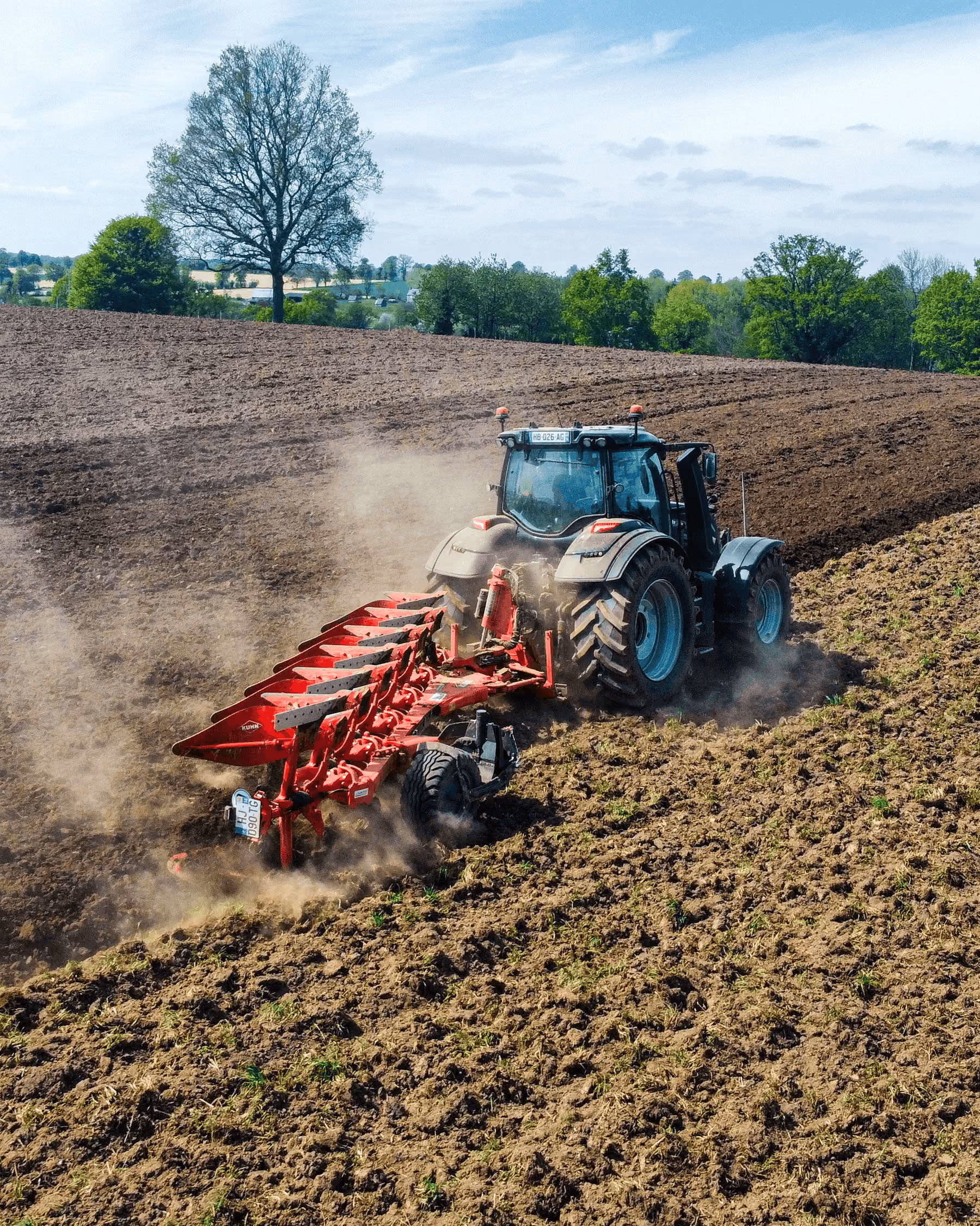 Tractor with large red plow cultivates a wide open field under a partly cloudy sky with trees in the background.