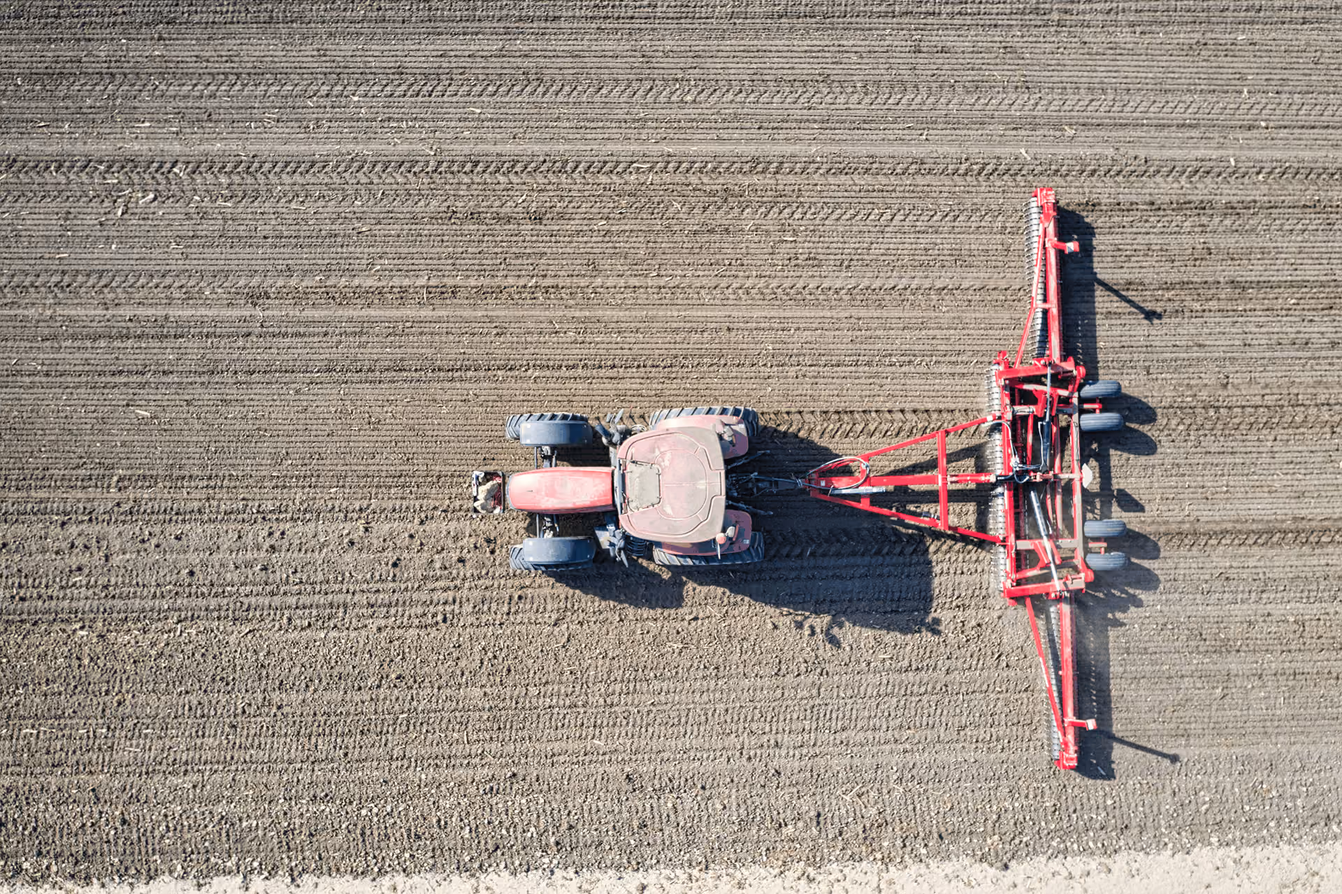 Aerial view of a red tractor pulling a wide cultivator across a plowed field, creating evenly spaced parallel furrows in the soil.