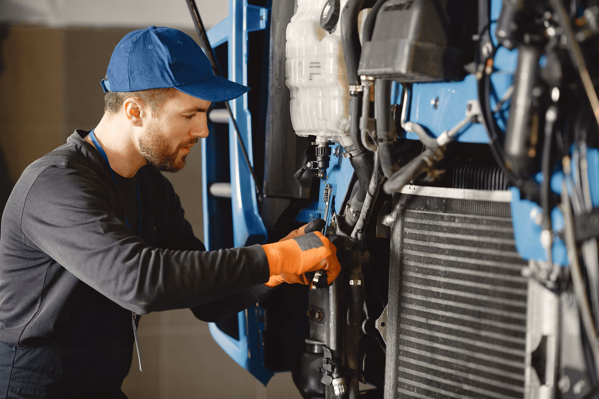 Mechanic wearing orange gloves and a blue cap uses a wrench to work on the front section of a large blue vehicle engine.