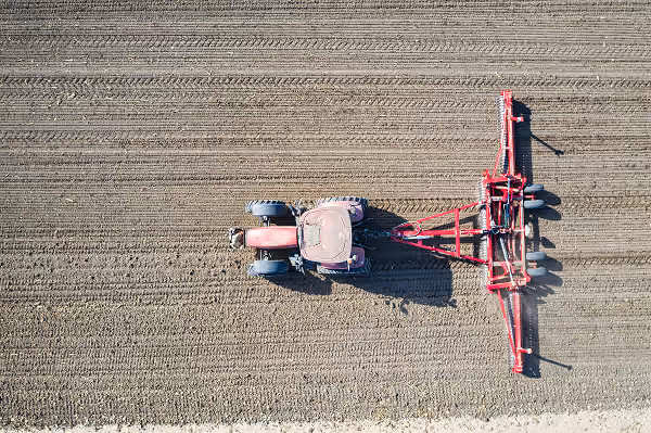 Aerial view of a red tractor plowing a field with evenly spaced furrows in dry soil.
