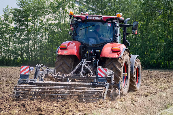 Red tractor tilling a plowed field with mechanical equipment attached at the rear, surrounded by green trees in the background.